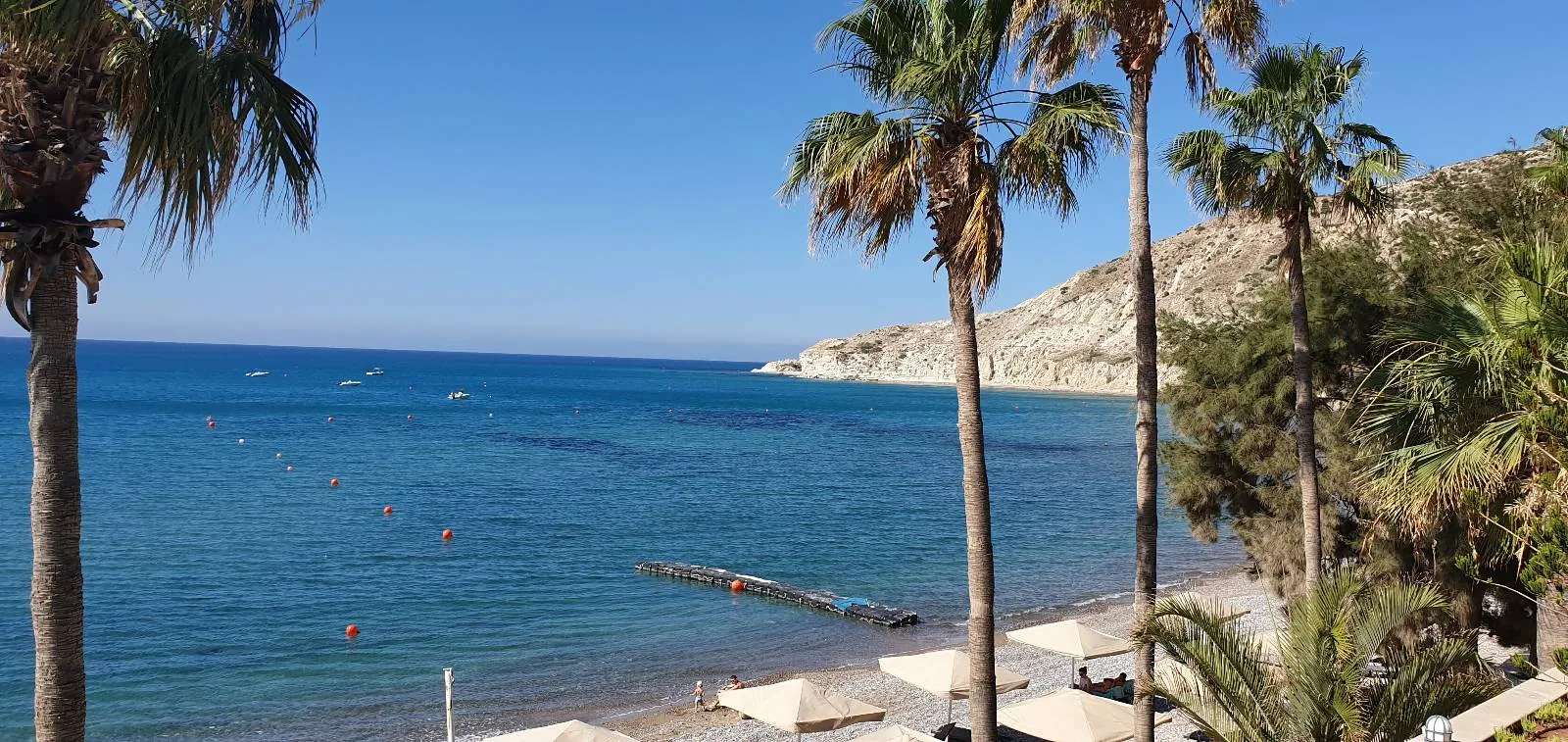 A scenic beach with clear blue water, palm trees, white sun loungers, and rocky hills in the background under a bright, sunny sky.