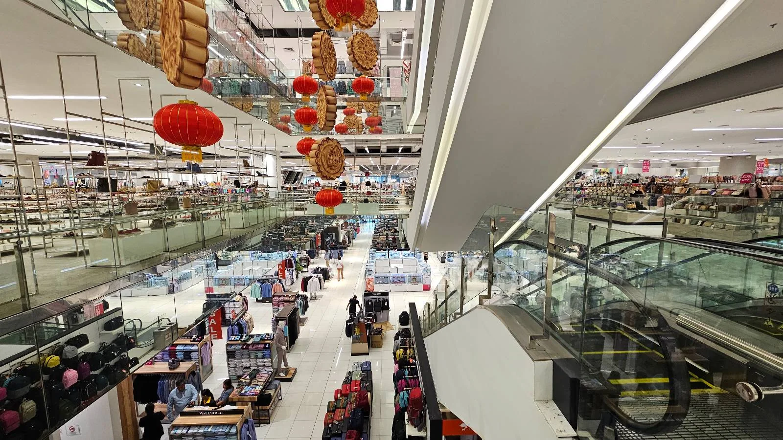 A large, bustling department store with multiple levels. Red lanterns are hanging from the ceiling as decorations. People are shopping, and escalators connect the floors. Shelves and displays are filled with various products.