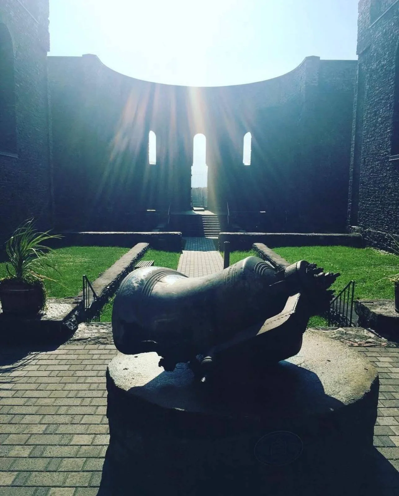 A bronze damaged bell is displayed in the open remains of a church with green grass, surrounded by stone walls and large windows letting in sunlight.