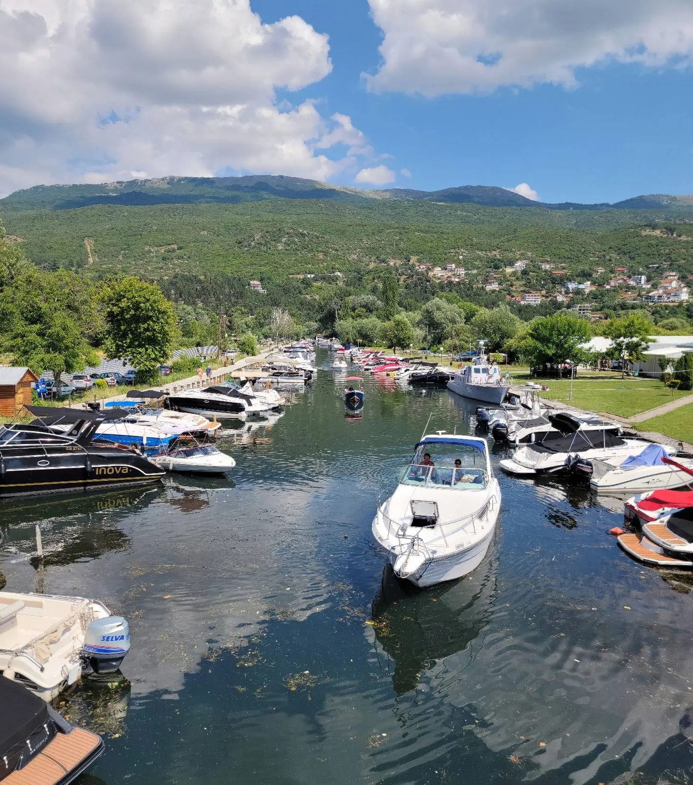 A scenic view of a marina with several boats docked along the water. A white boat is heading towards the open water. Lush green hills and a partly cloudy blue sky form the backdrop.