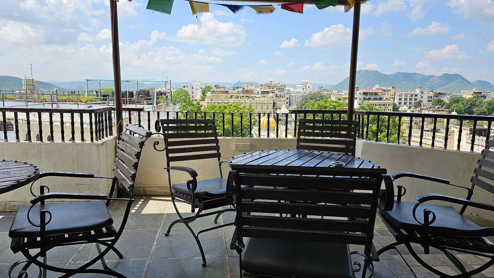 Outdoor patio with black metal table and chairs under a canopy, colorful prayer flags overhead, and a scenic cityscape with mountains in the background under a blue sky with clouds.