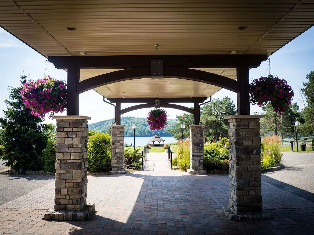 Covered stone walkway with hanging flower baskets leads to a scenic view of trees and hills under a clear blue sky.
