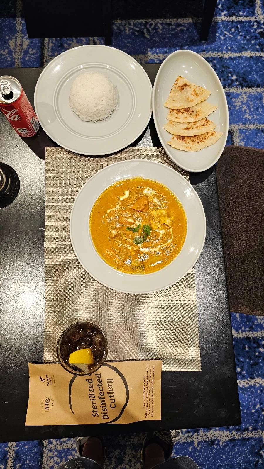 Overhead view of a meal with a plate of curry garnished with cream and herbs, a bowl of white rice, flatbread slices, a can of Coke, a glass of soda with lemon, and a brown paper napkin on a table.
