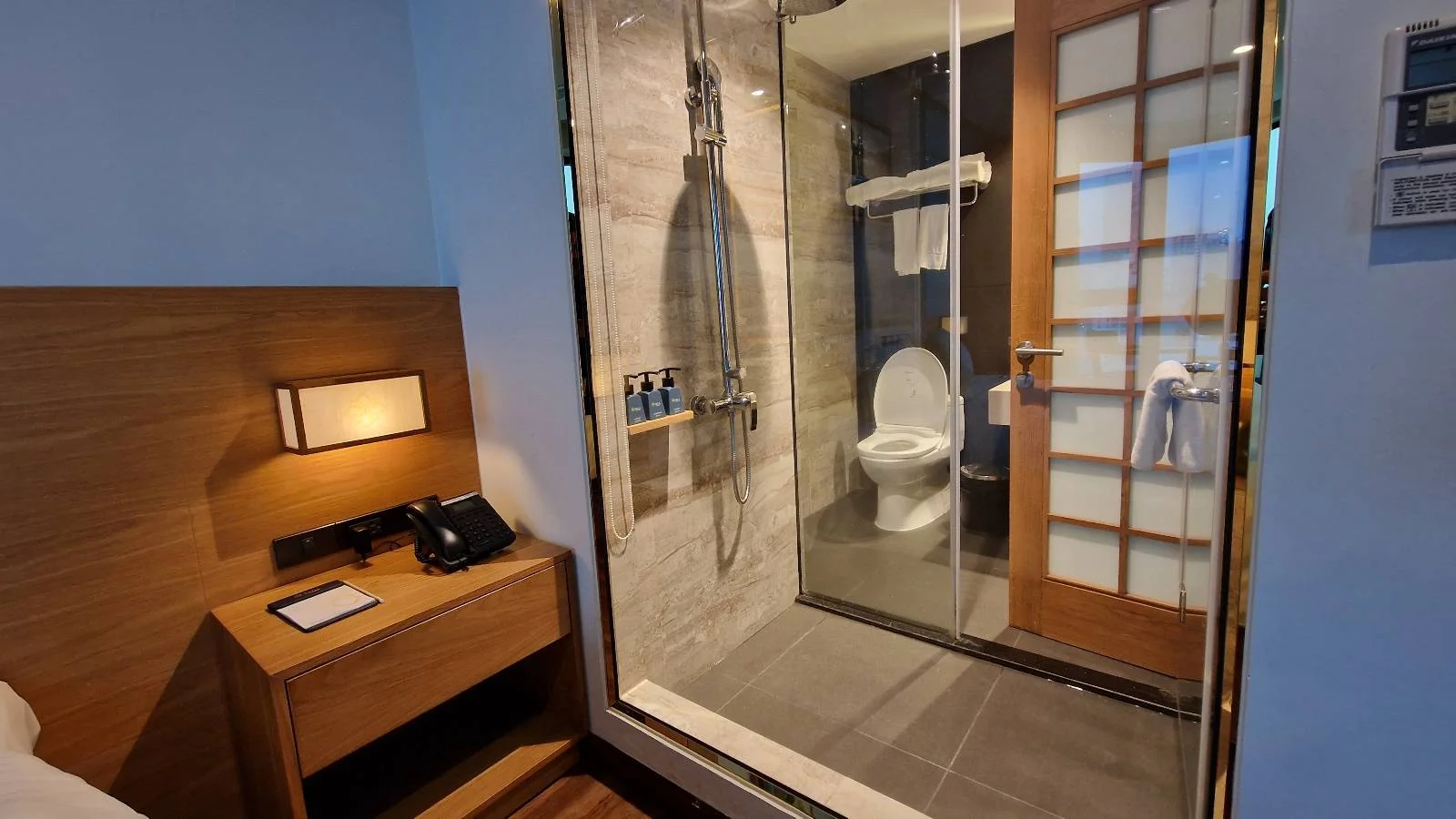 A hotel room with a view into the bathroom. The bathroom has a glass shower enclosure and a toilet. A bed, nightstand with a phone, lamp, and tablet are visible in the foreground, separated by a glass wall with a sliding wooden door.
