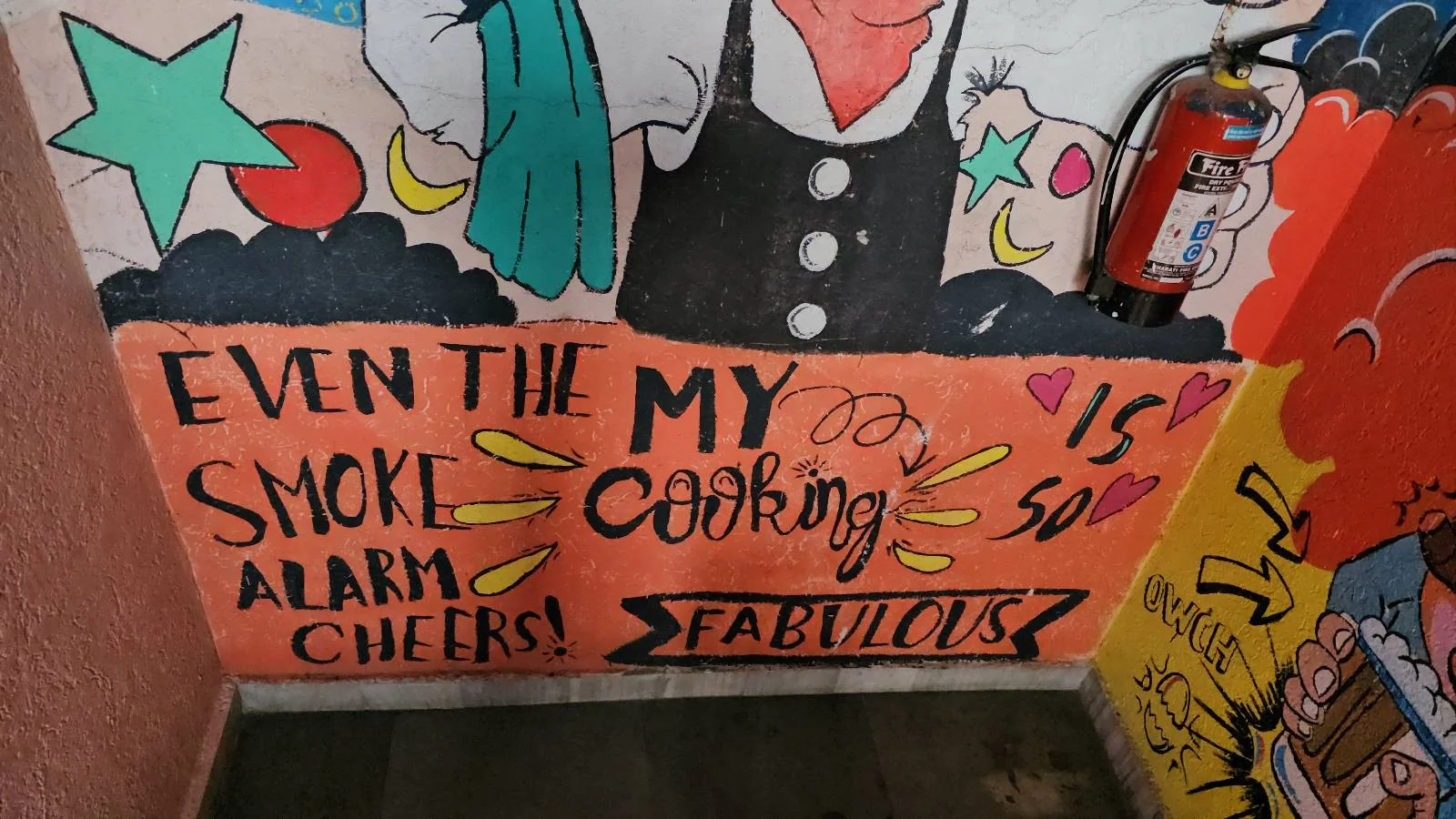 A colorful mural on a wall features the text "Even the smoke alarm cheers! My cooking is so fabulous." The background includes stars, moons, and a fire extinguisher on the right side.