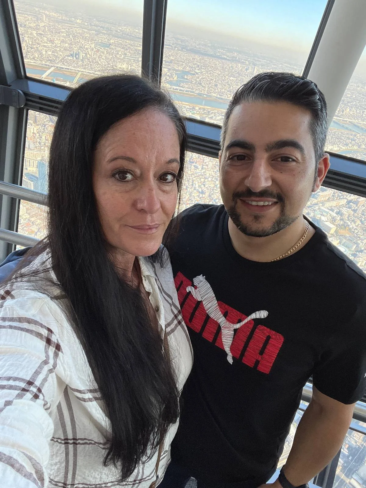 A woman and a man smiling for a selfie inside a tall building with large windows. The cityscape is visible in the background. The woman has long dark hair and is wearing a white shirt, while the man has short dark hair and a black Puma T-shirt.