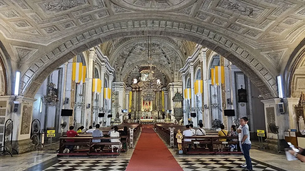 Interior of a grand cathedral with ornate ceiling, chandeliers, and yellow banners. People sit on pews, a red carpet leads to the altar.