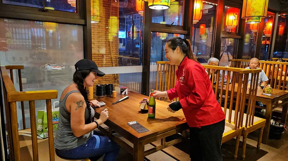 Woman in a baseball cap smiles as server in red jacket presents a drink at a wooden table. Warmly lit restaurant with red lanterns.
