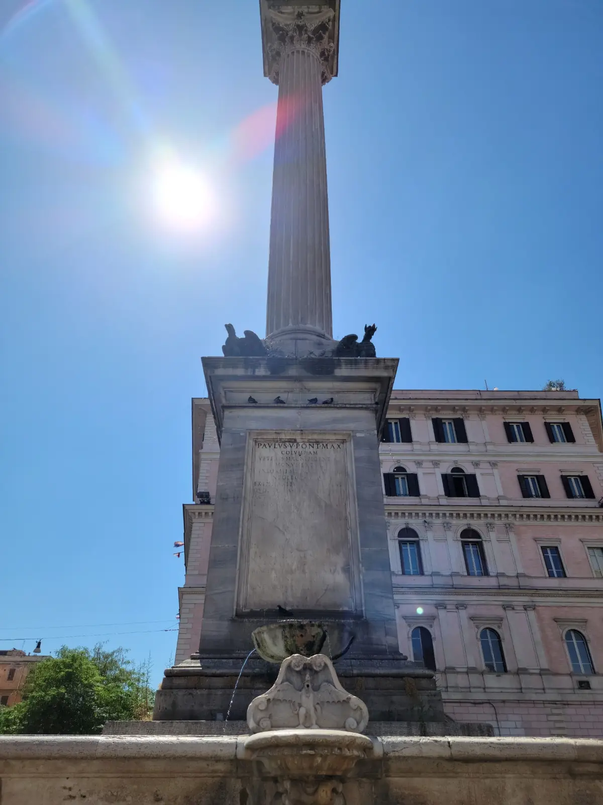 A tall stone column with a lion statue at its base stands in front of a multi-story building under a bright blue sky and sunlight.