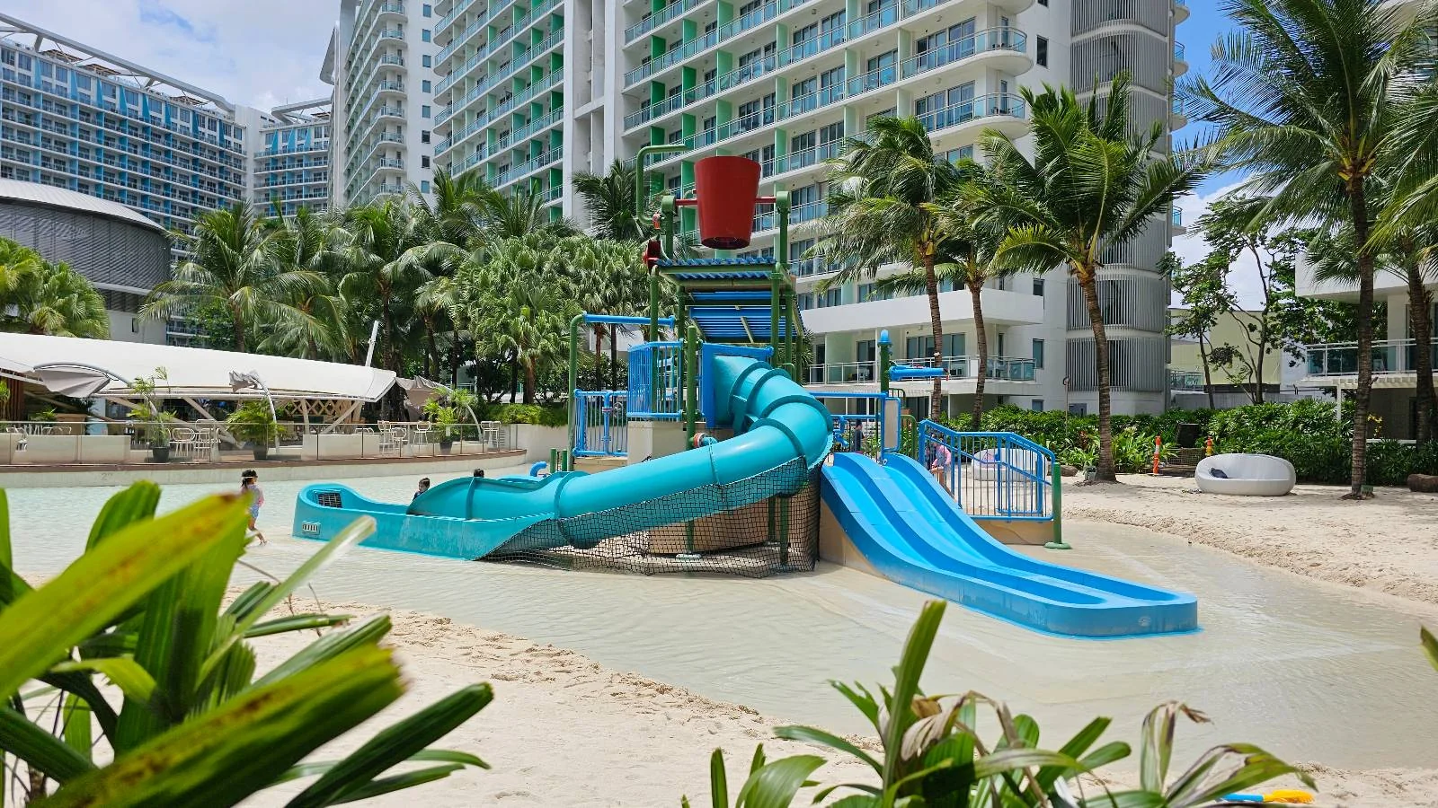 A small water playground with blue slides and a red water bucket, surrounded by palm trees and plants, is set in front of a modern multi-story hotel or apartment building.