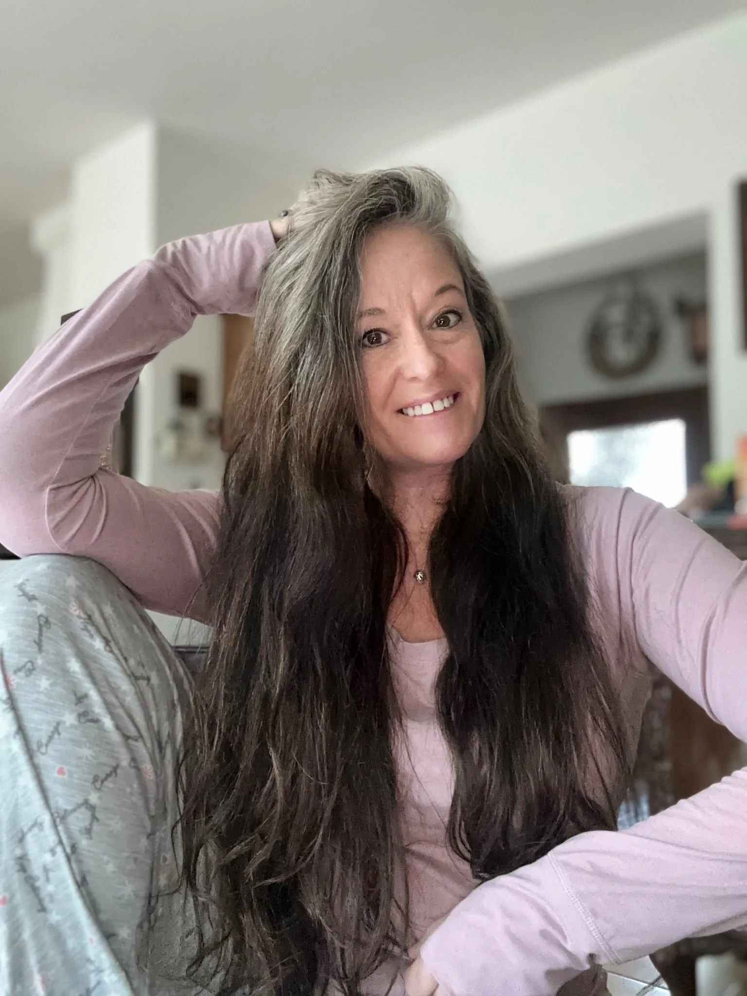 A close-up selfie of Jenn smiling warmly at home, running her hand through her long hair.