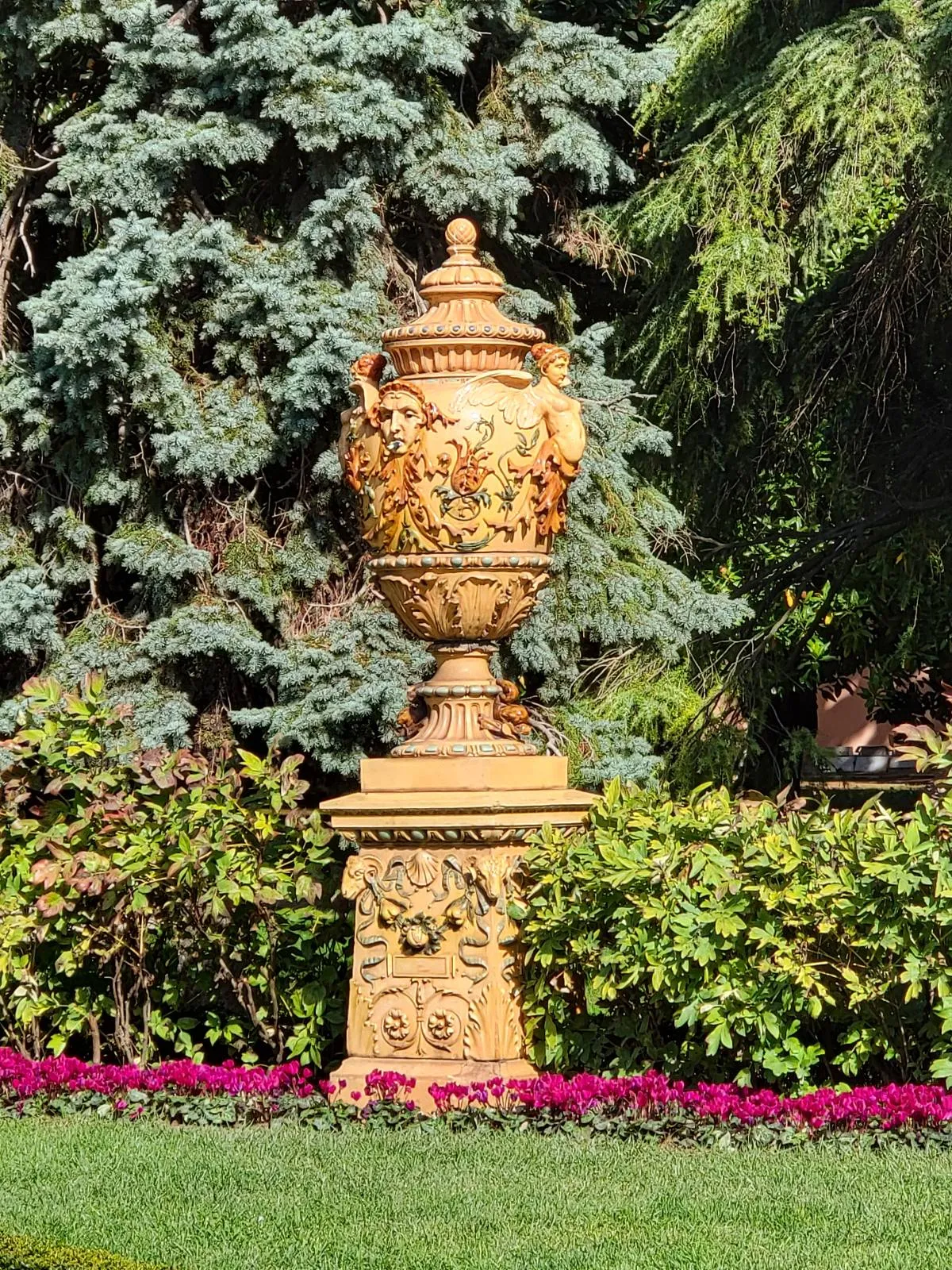 A decorative stone urn with detailed carvings stands on a pedestal in a garden, surrounded by green foliage and pink flowers, with trees in the background.