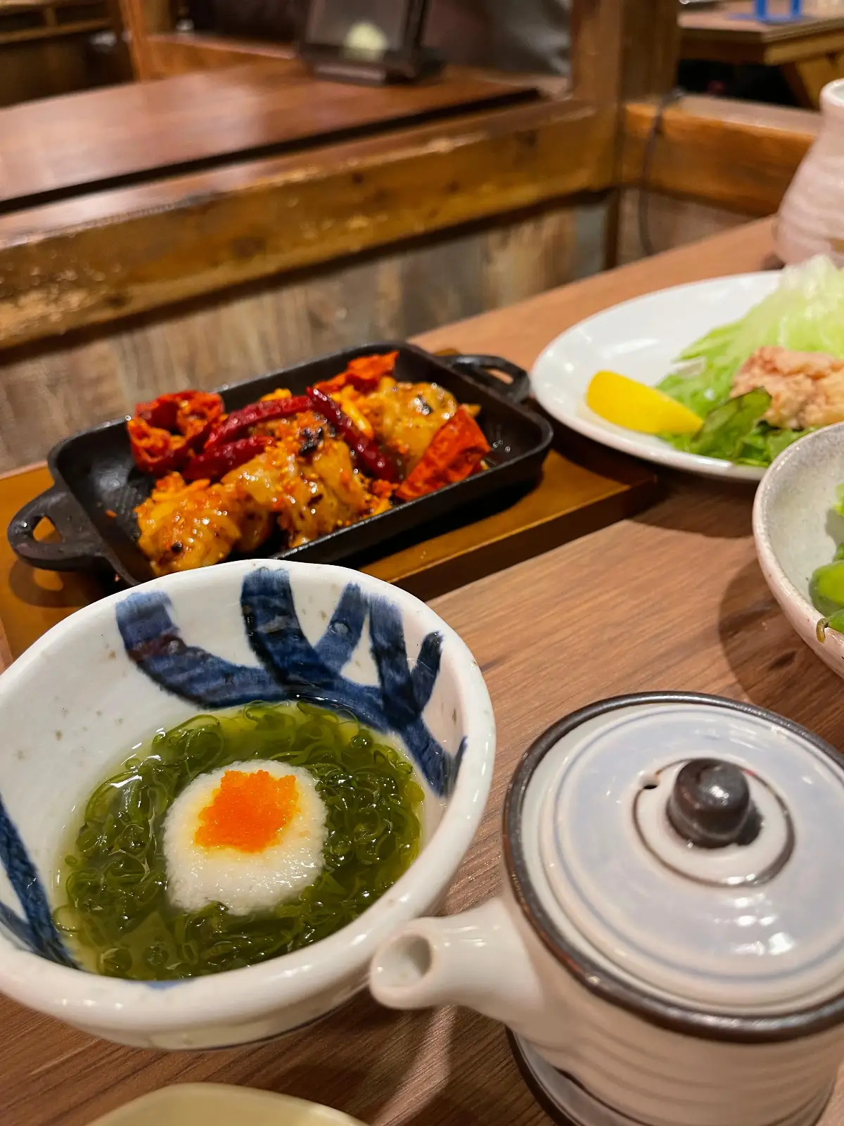 A table set with Japanese dishes, including a bowl with seaweed and a raw egg yolk, a plate of cooked meat and vegetables, and a side of salad with yellow pickles.