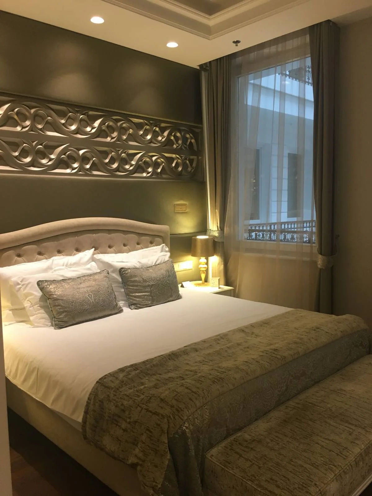 A cozy bedroom with a neatly made bed featuring a white duvet, gray pillows, and a green throw. The walls are dark with a decorative panel, and there's a window with sheer curtains. A bedside lamp emits a warm glow.