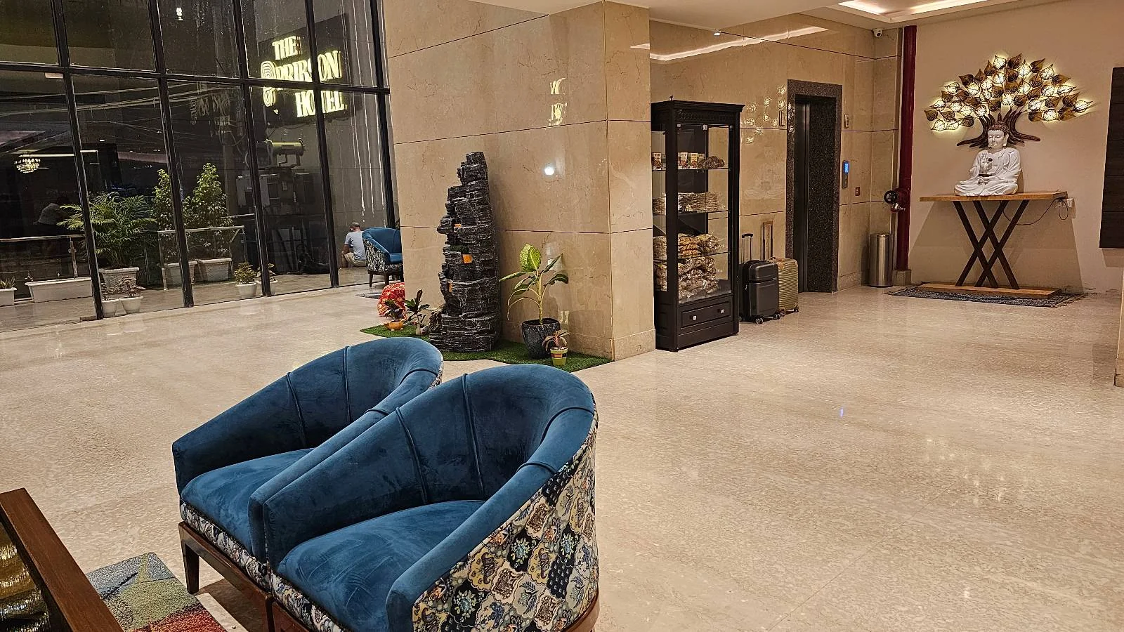 Hotel lobby with two blue velvet chairs, a glass display cabinet, plants, and a decorative statue. Large floor-to-ceiling windows and a table with a floral arrangement are visible in the background.