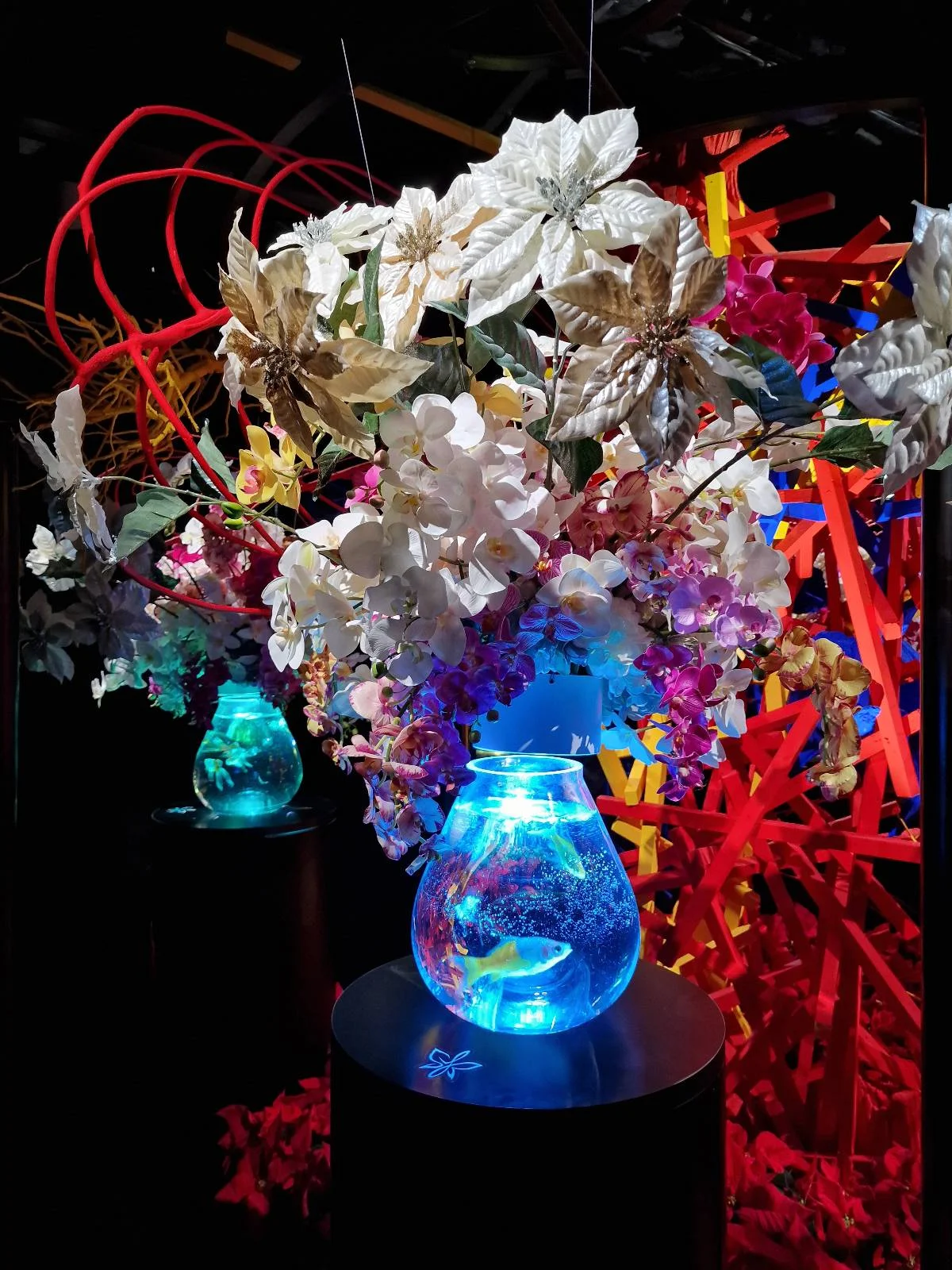 A vibrant floral arrangement with white and pink flowers is illuminated by a glowing blue vase. Behind it, an abstract backdrop of red and orange elements adds a dramatic contrast, enhancing the vividness of the scene.