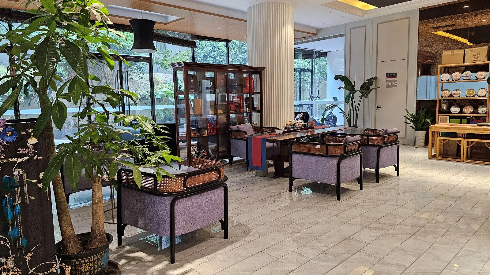A bright and modern tea room with wooden tables and chairs, large windows, potted plants, a glass display cabinet, and shelves with teapots and cups. Natural light fills the clean, spacious interior.