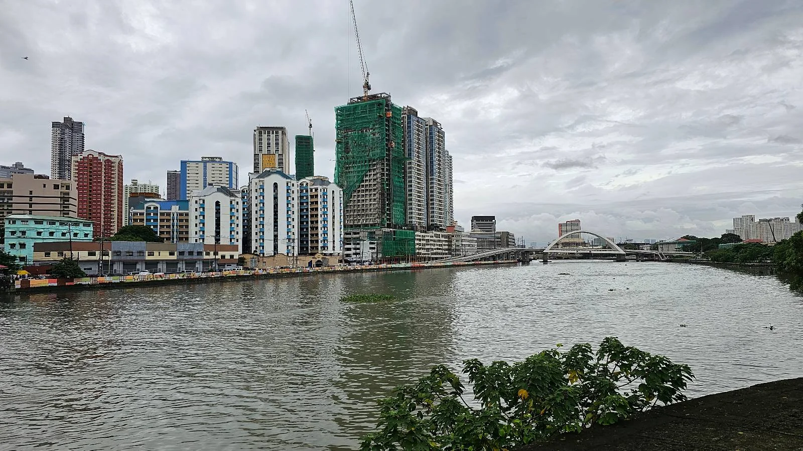 A river flows through a cityscape under a cloudy sky. Tall buildings, some under construction, line the riverbank. Lush green foliage is visible in the foreground, adding contrast to the urban scene.