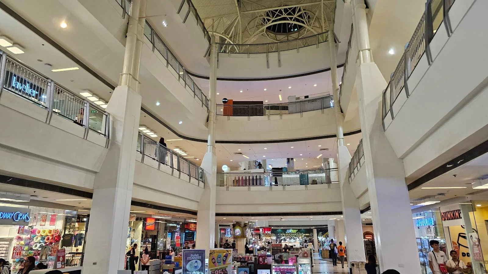 A multi-story shopping mall with several shops visible on the ground floor. People are walking and browsing. The ceiling features a circular design, and there are railings on each level. Bright lighting illuminates the interior.