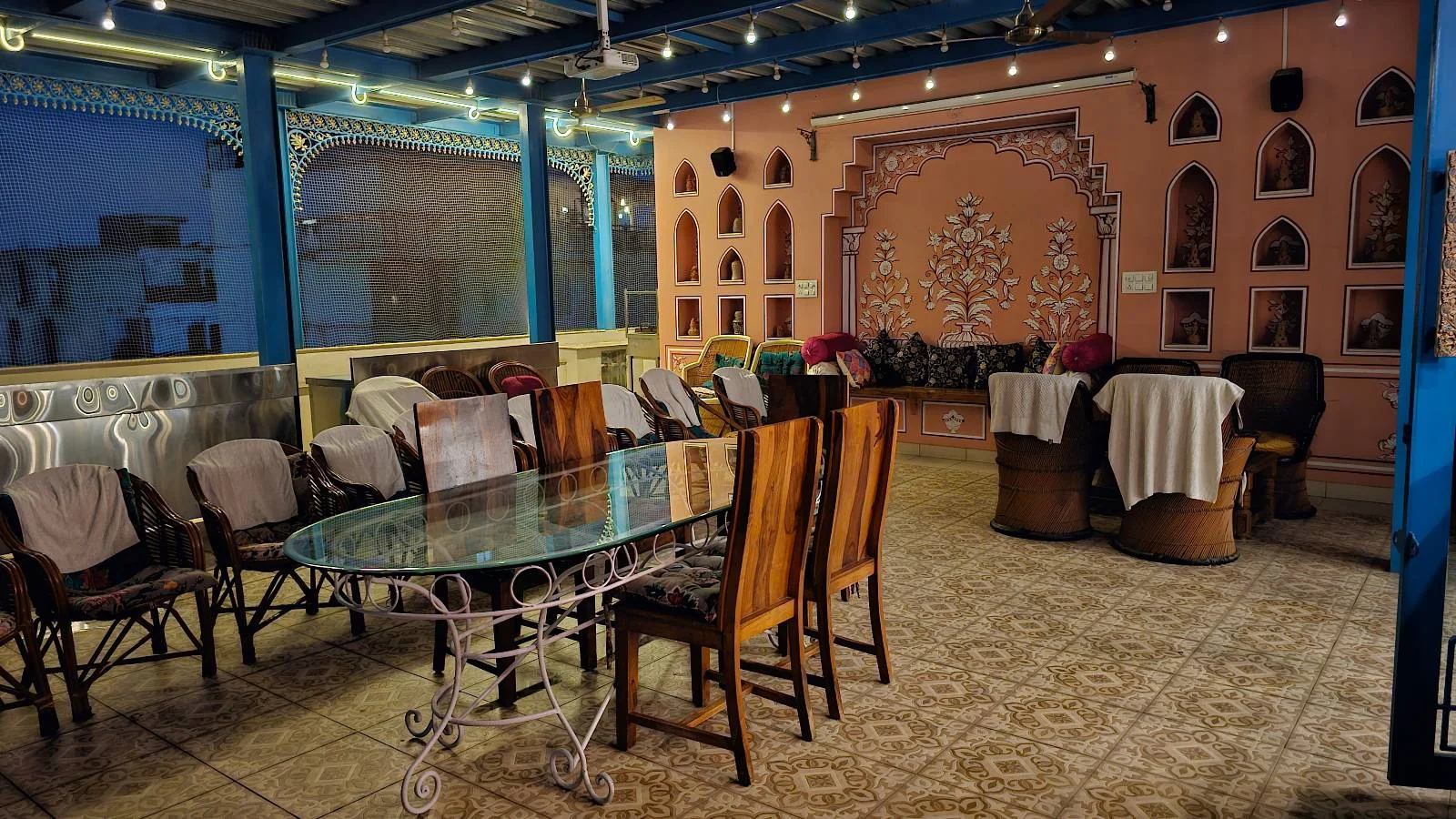 A cozy restaurant interior with blue accents, a glass-top table with wooden chairs, wicker armchairs, and decorative wall niches. String lights hang from the ceiling, creating a warm, inviting atmosphere.