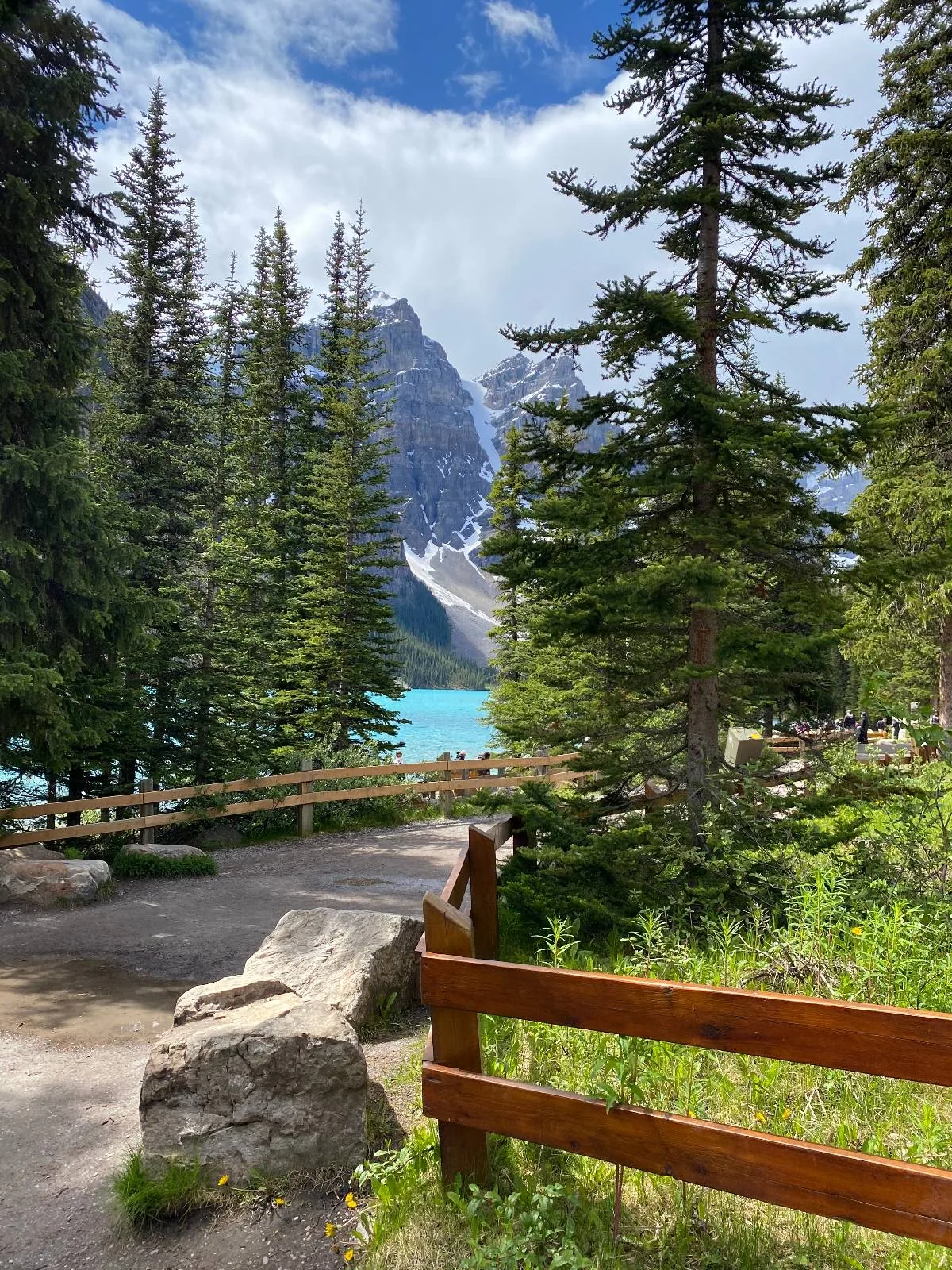 A scenic pathway lined with pine trees and wooden fencing leads to a turquoise lake, with a rocky mountain peak rising in the background under a partly cloudy sky.