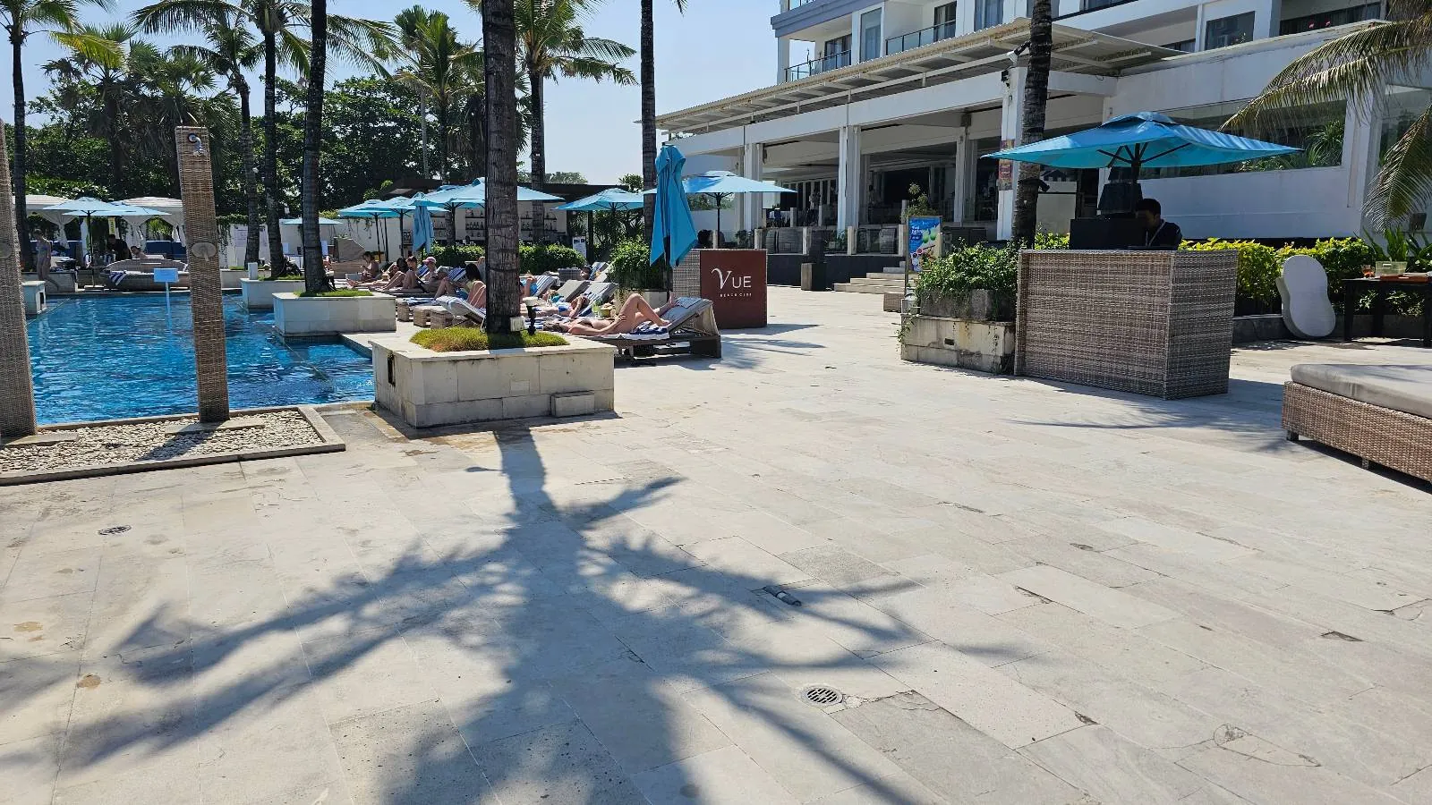Sunlit poolside area at a resort, with lounge chairs and umbrellas beside a clear blue pool. Palm trees cast shadows on the paved ground. A building with a shaded patio is in the background.