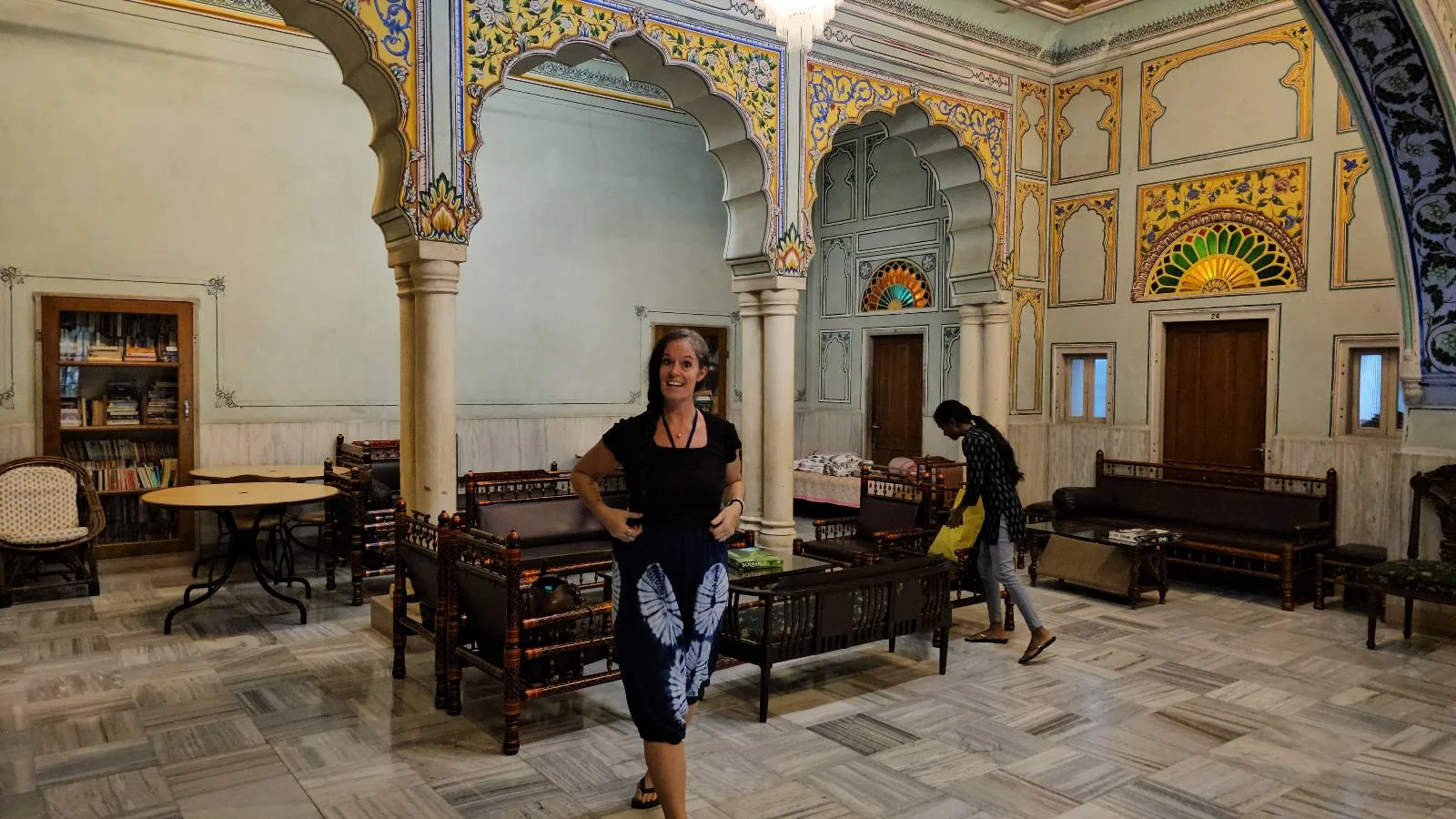 A woman stands in the center of an ornately decorated room with arches, stained glass windows, tables, and chairs, while another person arranges items on the right side. The room has intricate designs and a marble floor.