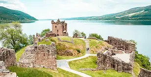 Ruins of Urquhart Castle overlooking Loch Ness in the Scottish Highlands, showing stone walls, towers, and footpaths set against the calm freshwater loch and surrounding hills.