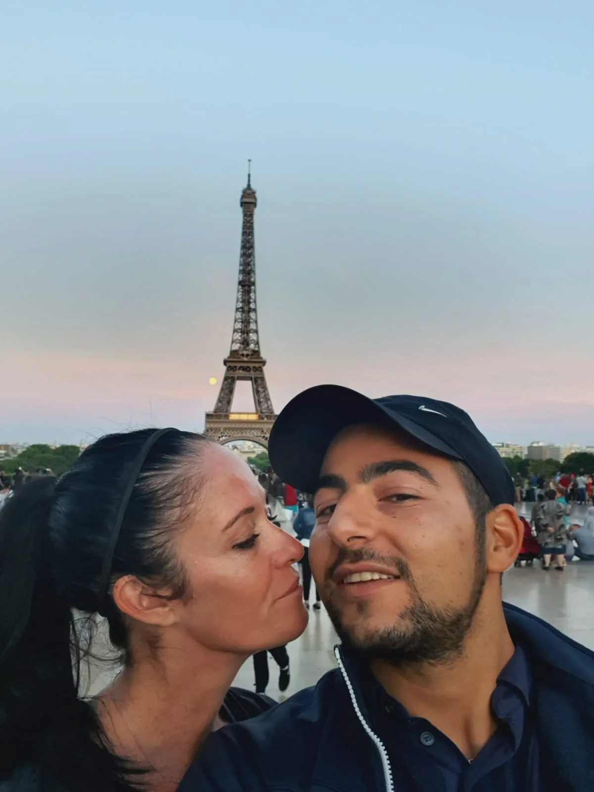 A couple taking a selfie with the Eiffel Tower in the background. The woman is kissing the man's cheek.