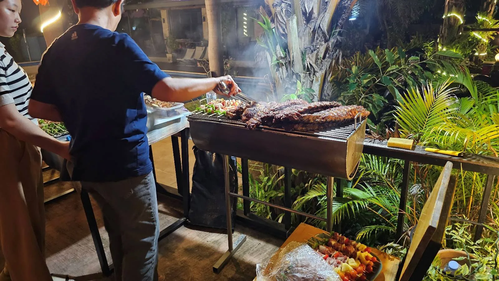 Two people grilling meat and vegetables on an outdoor barbecue at night, with foliage in the background.