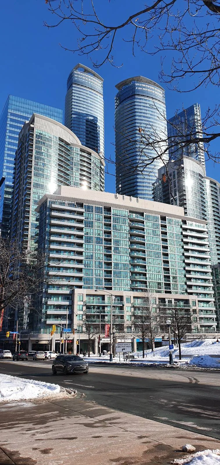 Skyscrapers with reflective glass in a city on a clear, sunny day. Cars drive on a snowy street. Bare tree branches frame the scene.