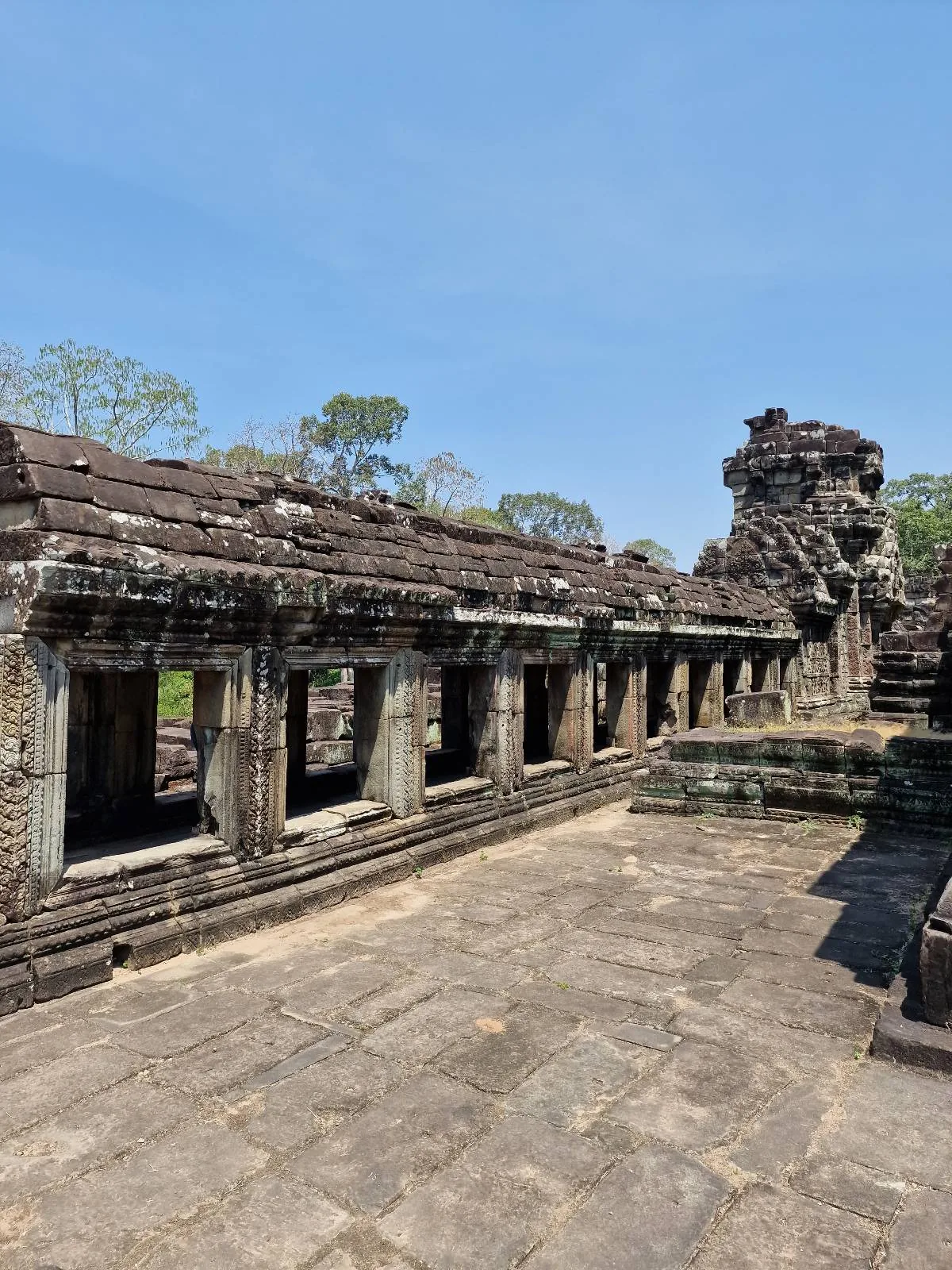 Ancient stone ruins with columns and carvings under a clear blue sky, surrounded by sparse trees and greenery, likely part of a historic temple or archaeological site.