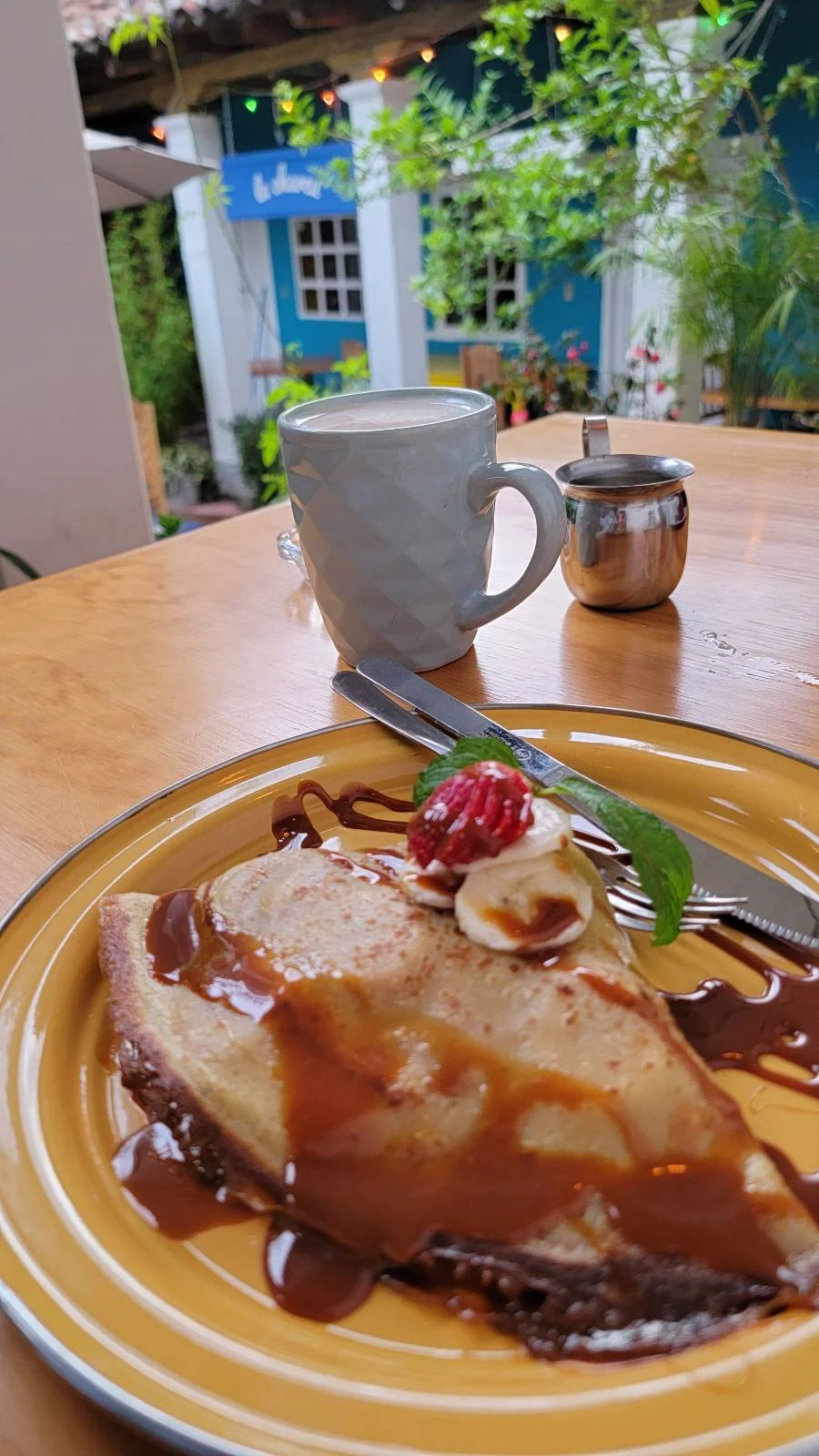 A dessert crepe topped with banana slices, a raspberry, mint leaves, and drizzled with chocolate sauce sits on a yellow plate. A cup of coffee and a small pot of milk are on the wooden table. A garden is visible in the background.