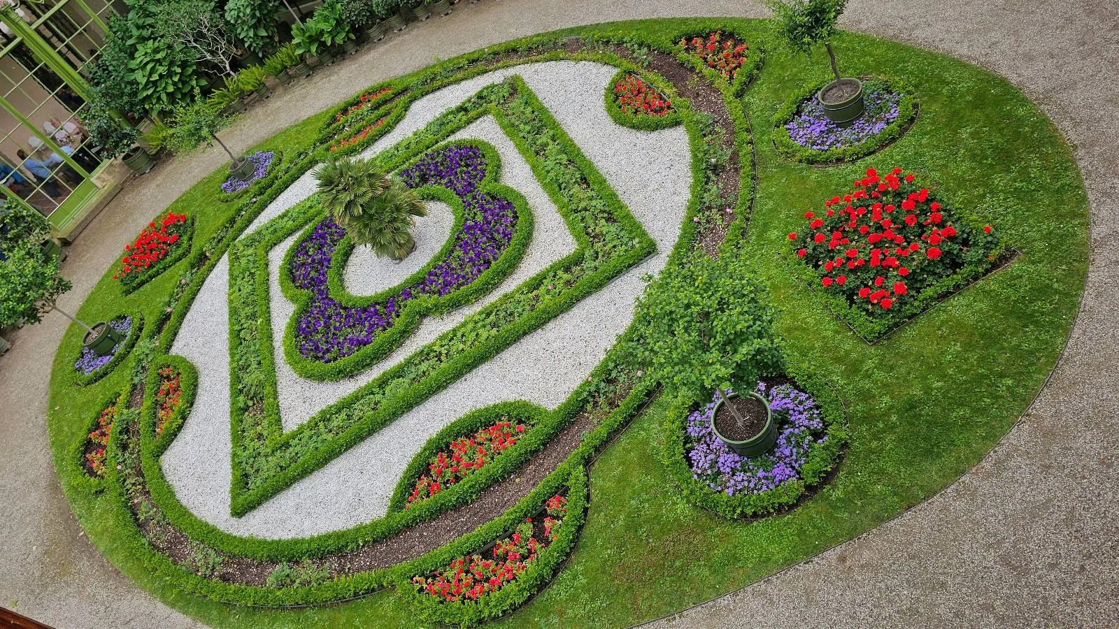 An ornate circular garden features geometric flower beds with colorful red, purple, and green plants arranged in symmetrical patterns, all bordered by neatly trimmed grass paths.