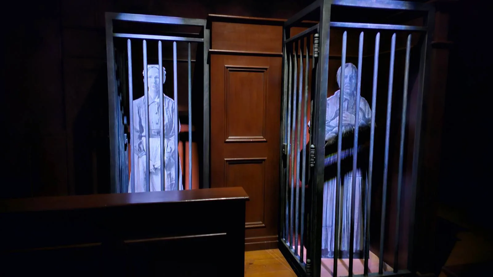 Two life-size statues stand in separate jail cells with metal bars, illuminated by overhead light. A wooden wall and a bench or partition are in the foreground, creating a courtroom or prison-like scene.
