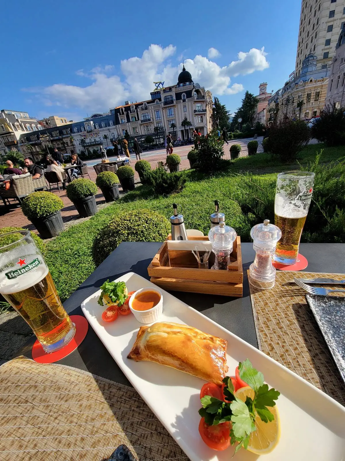 A plate with a pastry, salad, and sauce sits on an outdoor café table with two glasses of beer. In the background, a city square, greenery, and historic buildings are visible under a partly cloudy sky.