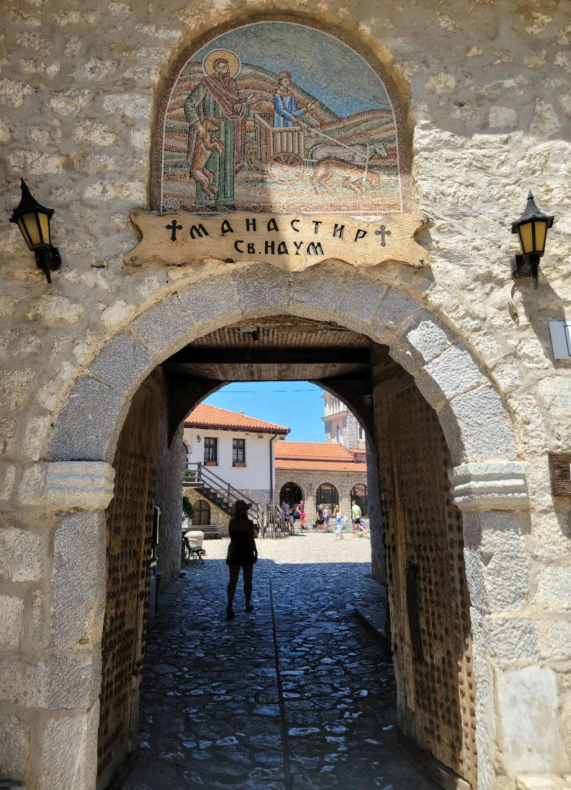 Stone entrance arch to a monastery with a mosaic depicting saints above. The wooden sign reads "ΜΑΝΑΣΤИРЪ СЬ. НАУМЪ." Lanterns are on either side. A cobblestone path leads to buildings in the background with a person walking.