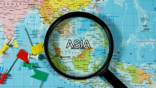 A magnifying glass hovers over a map of Asia, highlighting countries like China, India, and Indonesia. The word "ASIA" is bold and centered under the glass, with colorful push pins visible on surrounding areas of the map.