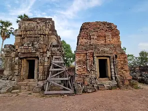 Ancient stone and brick ruins in Cambodia, with two weathered structures under a blue sky. Wooden scaffolding supports one building, while sparse trees in the background enhance the scene's historical atmosphere. This idyllic setting is a must-see on any travel guide without needing exhaustive visa information.