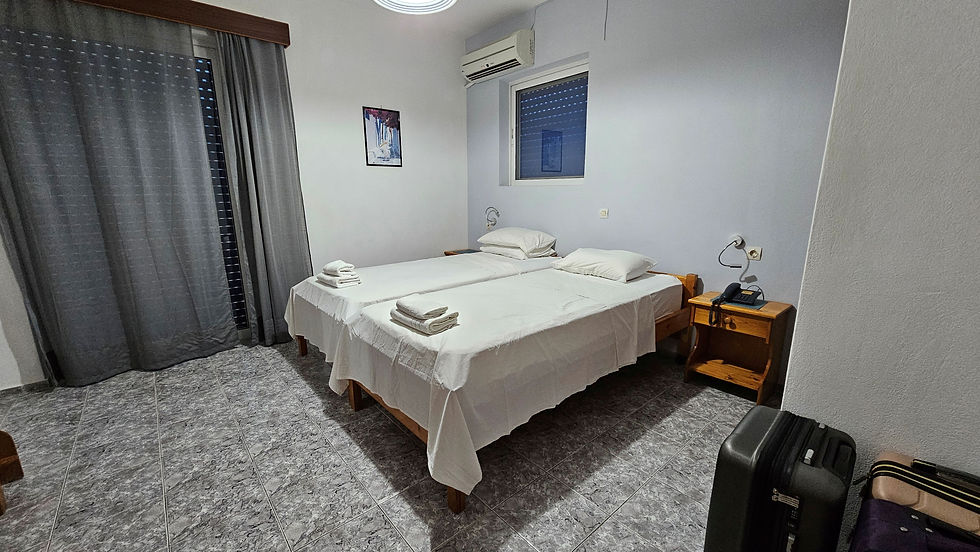 Two neatly made twin beds with white sheets in a tidy room. Gray curtains cover a window, and a suitcase is by the wall. Calm ambiance.