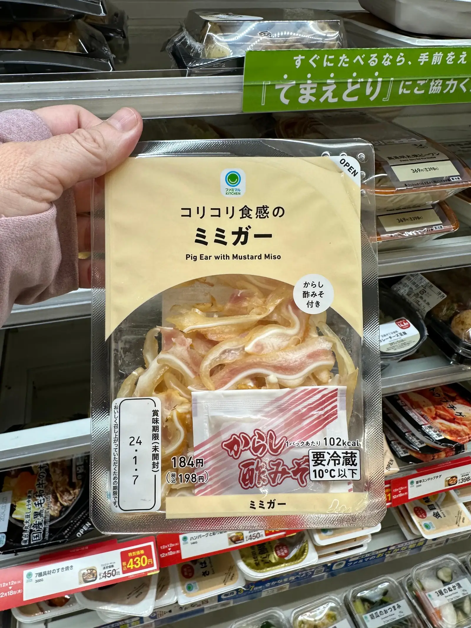 A hand holds a packaged tray of sliced pork ears with Japanese labeling in front of a supermarket refrigerated shelf displaying other food items.