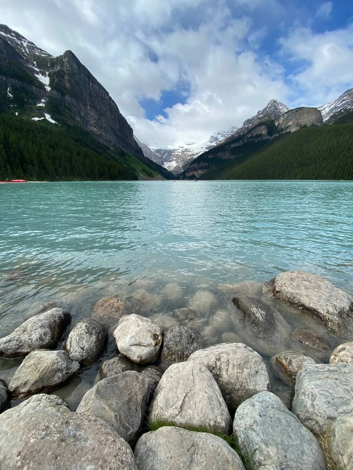 A clear turquoise lake with rocky shore in the foreground, surrounded by forested mountains and snow-capped peaks under a partly cloudy sky.