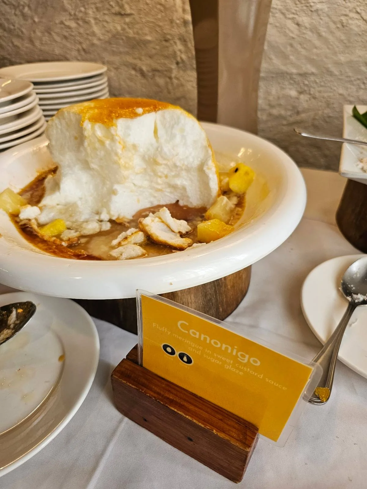 A dessert labeled "canonigo" with a fluffy texture, surrounded by small pineapple pieces and caramel sauce, is placed on a buffet table with plates and utensils nearby.
