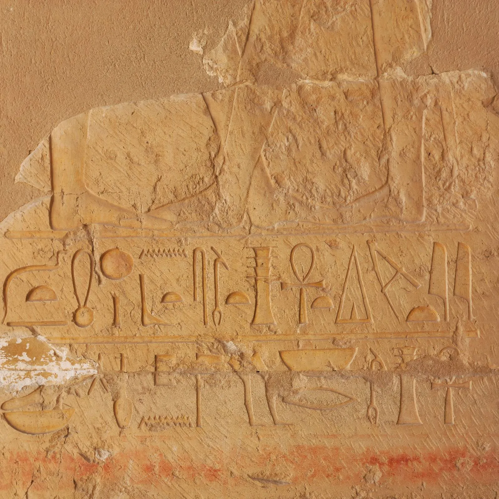 Ancient Egyptian hieroglyphs carved into a beige stone wall, featuring various symbols and shapes. The surface appears weathered and slightly faded with some red coloring visible near the bottom edge.