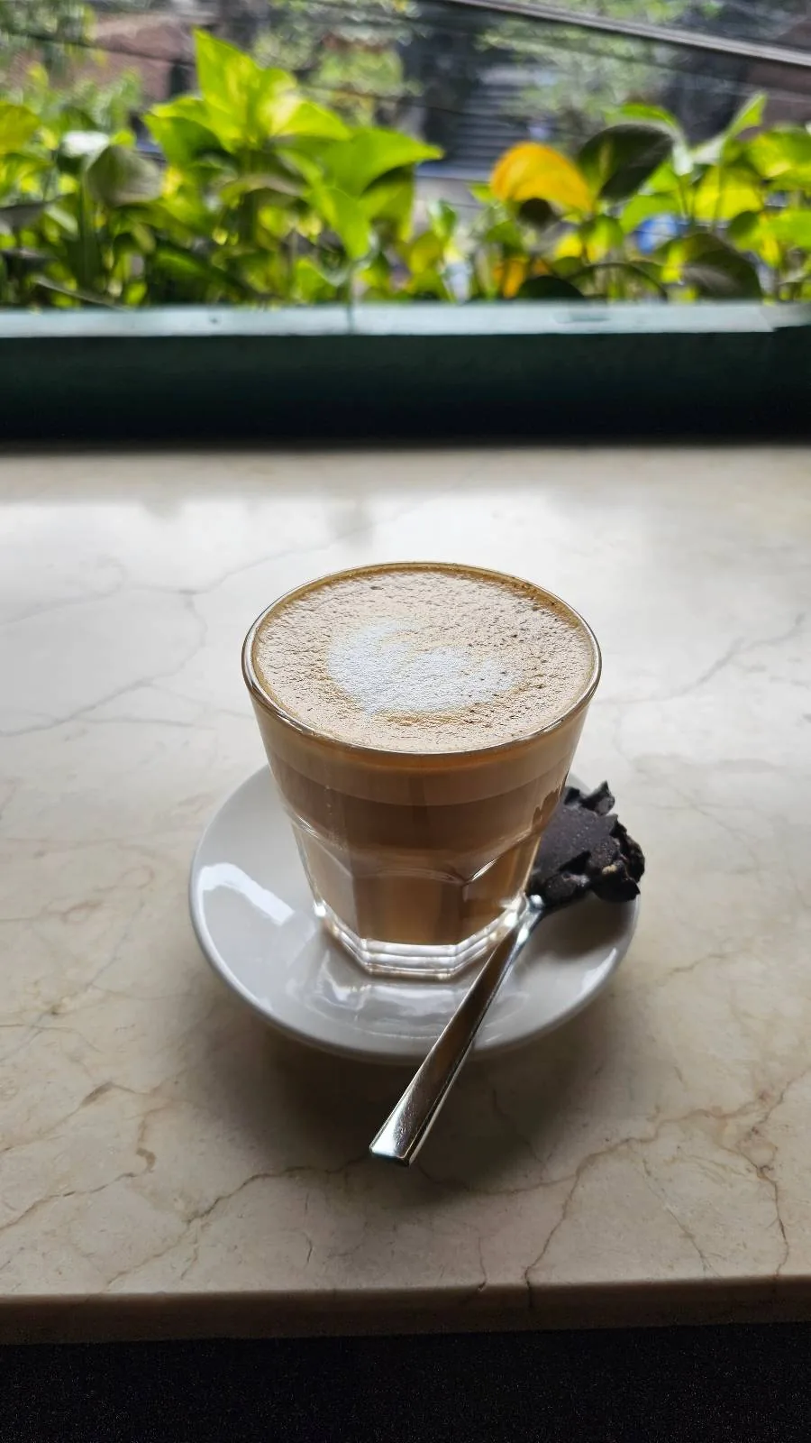 A glass cup of frothy coffee is placed on a white saucer with a silver spoon beside it. The setting is a marble table by a window with green plants visible outside.