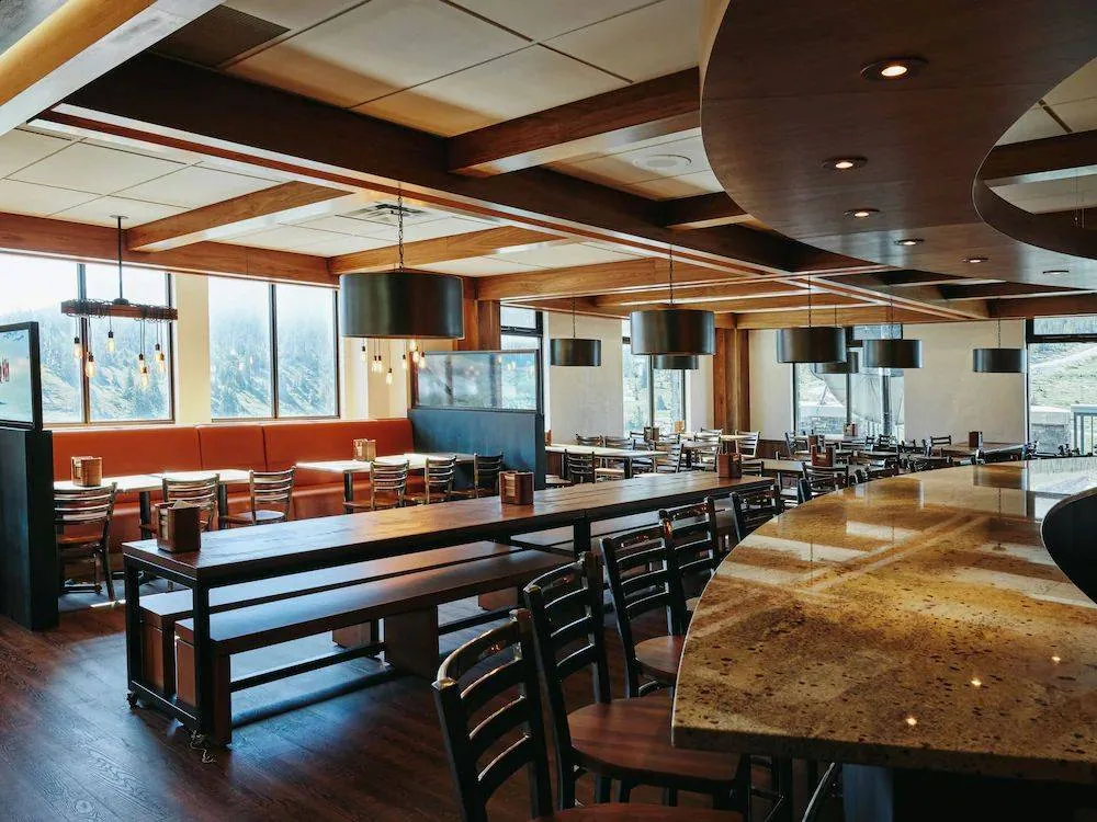 A modern, empty restaurant with wooden decor. The space features long wooden tables and chairs, large pendant lights, and a curved counter. Sunlight streams through large windows, illuminating the warm tones of the interior.