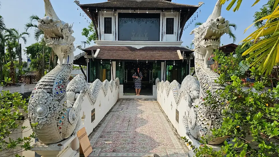 Ornate white statues flank a bridge leading to a building. A woman in black and white stands in the entrance. Trees and tiles enhance scene.