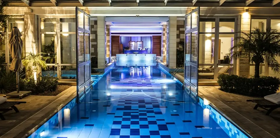 Illuminated indoor pool with blue tiles, flanked by loungers and palm plants. Open doors lead to a well-lit room, creating a serene atmosphere.