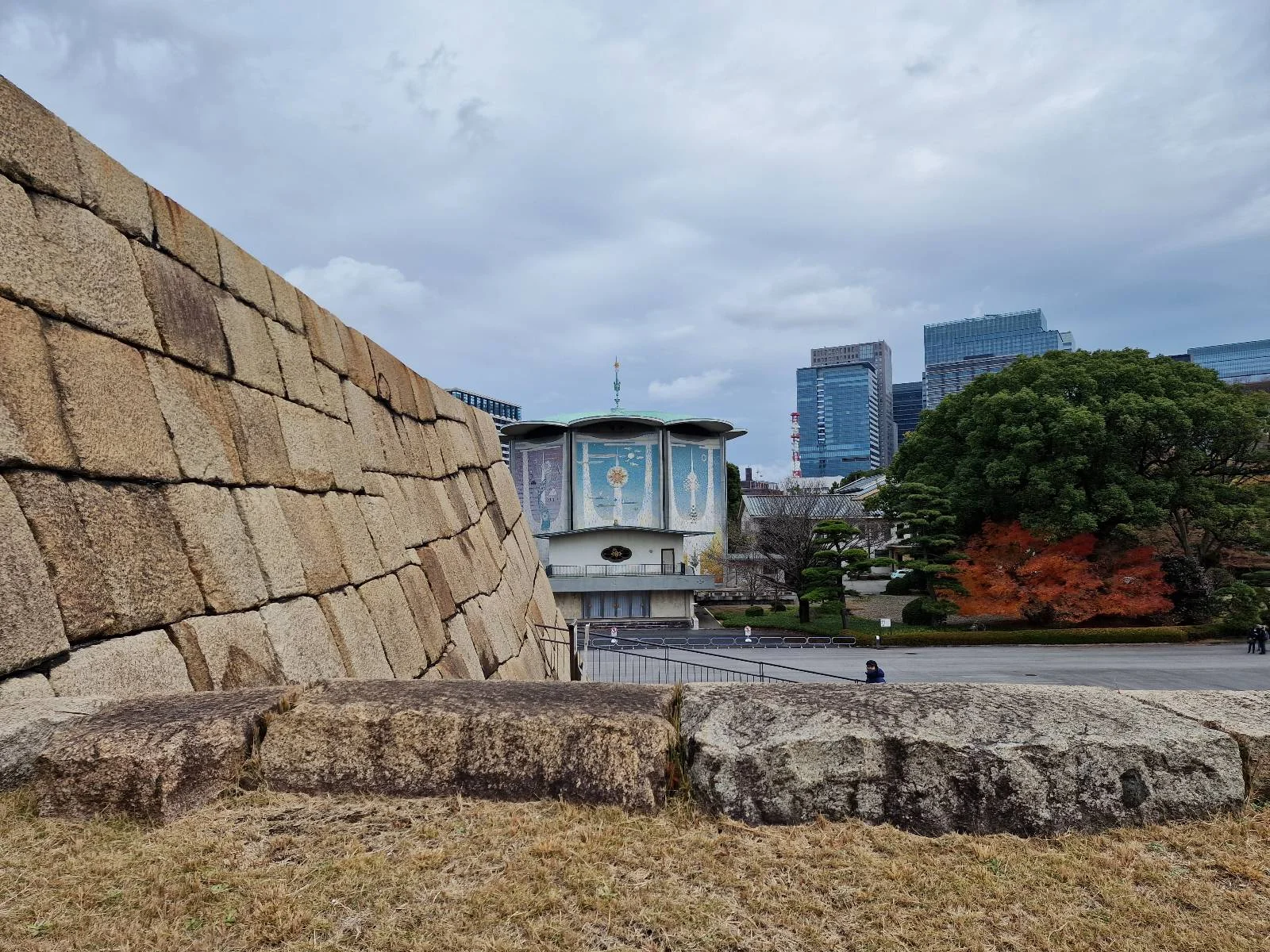 A large stone wall with a wide base is in the foreground. Behind it, a building with a mural and modern skyscrapers are visible. There's a large tree with orange leaves to the right under a cloudy sky.
