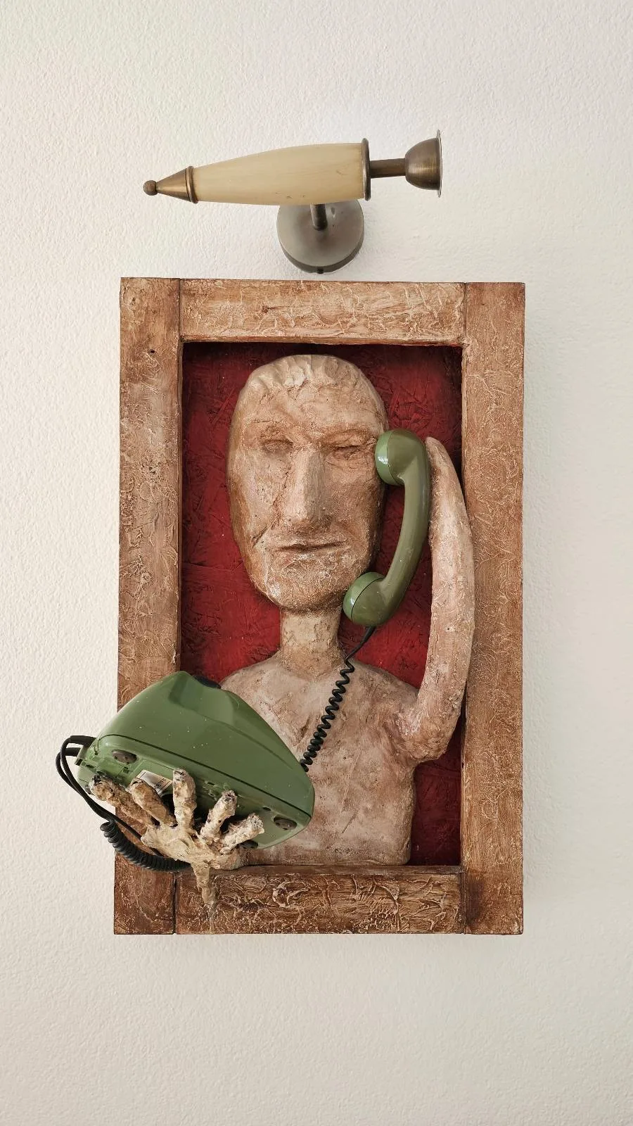 A wooden sculpture of a stylized human figure holding a green telephone receiver, set in a wooden frame with a red background; a green book and hand rest on the lower frame edge, and a cone-shaped object is mounted above.